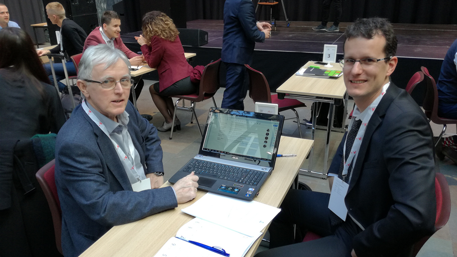 Two business men sitting by a table with a lap-top between themselves.