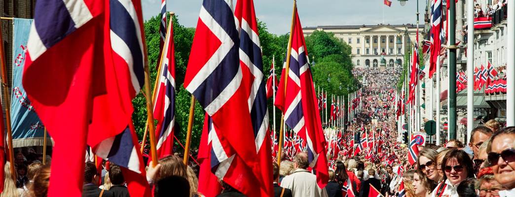 Foto com uma rua cheia de pessoas com bandeiras da Noruega. Ao fundo, tem a vista do Palácio Real. - Foto:Asgeir Helgestad_VisitNorway